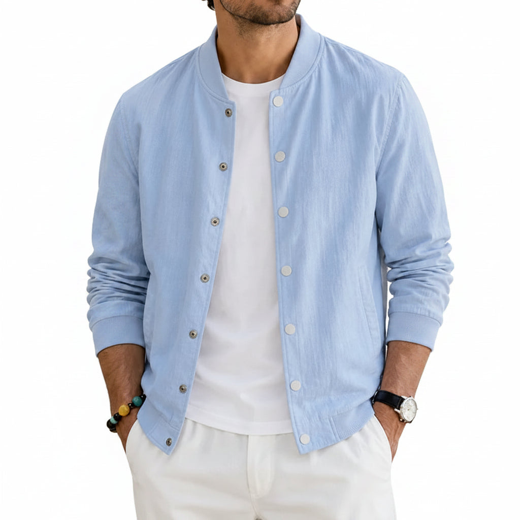 A man wearing a light blue button-up linen-blend jacket over a white t-shirt and white pants, standing in a sunlit outdoor patio setting.